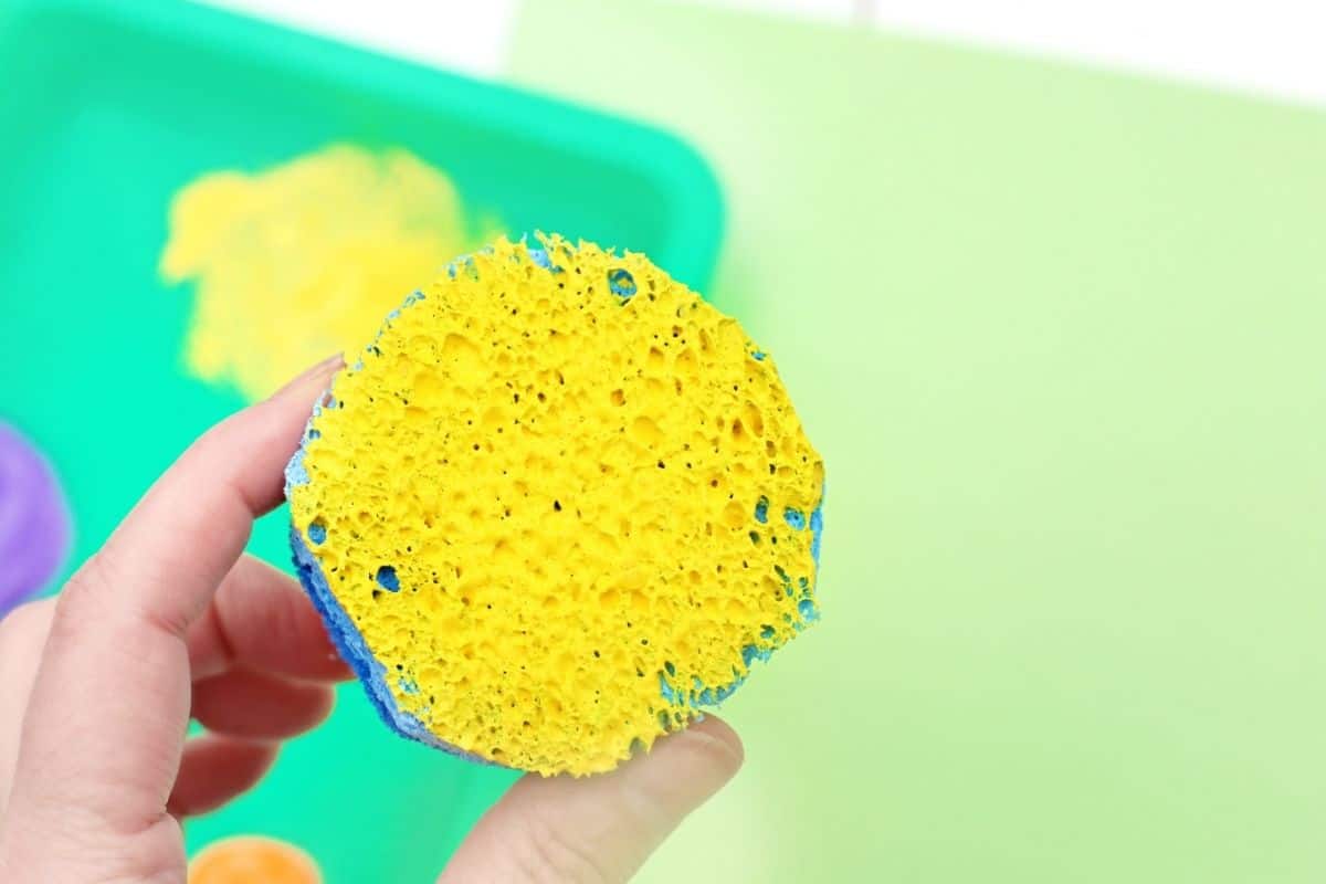 Hand holding a round sponge covered in bright yellow paint above green paper for a sponge painted caterpillar craft. The textured sponge is ready to stamp paint circles for the caterpillar art activity.