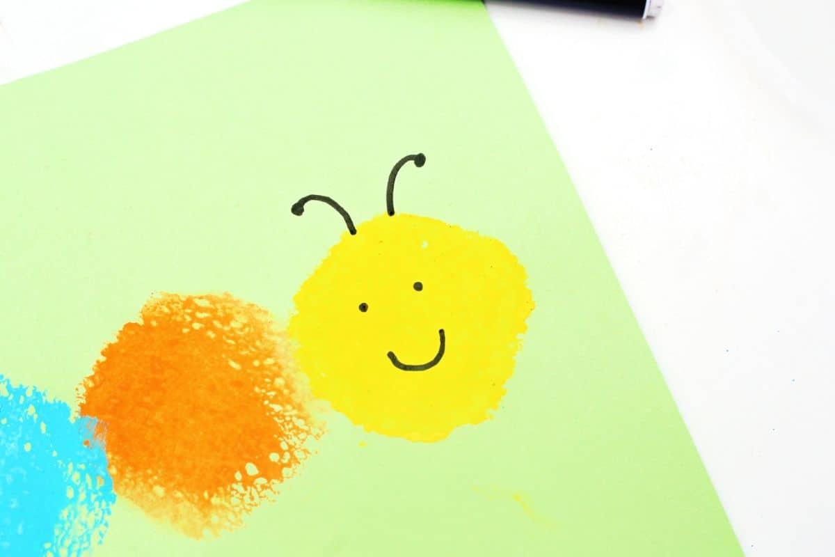 Close up of a sponge painted caterpillar head with a smiling face and black antennae on light green paper. The yellow sponge print sits beside orange and blue paint circles as part of the colorful caterpillar craft.