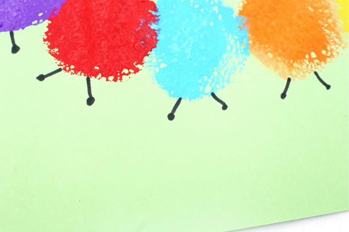 Close up of a sponge painted caterpillar body made with purple, red, blue, and orange paint circles on light green paper. Small black legs are drawn beneath each colorful sponge print to complete the caterpillar craft.