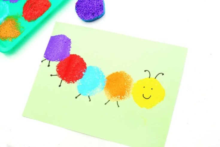 Sponge painted caterpillar craft made with purple, red, blue, orange, and yellow paint prints on light green paper. The yellow caterpillar head has a smiling face and black antennae, while paint covered sponges and a tray are visible in the background.