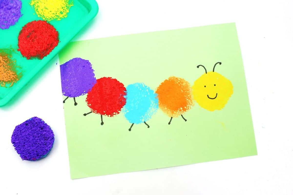 Finished sponge painted caterpillar craft on light green paper with purple, red, blue, orange, and yellow sponge print circles forming the caterpillar body. The yellow head has a smiling face and black antennae, with paint covered sponges and a paint tray placed beside the colorful bug themed activity.