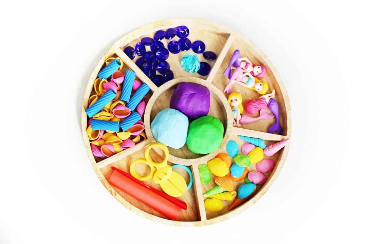 Round wooden tray filled with mermaid playdough activity materials including colorful shells, blue gems, mermaid figurines, playdough in purple, blue, and green, toy scissors, and playdough tools. The organized sensory play setup provides loose parts for creating underwater mermaid scenes.