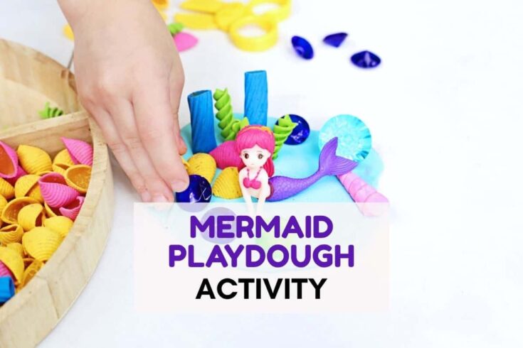 Child's hand arranging colorful shells, gems, and a pink haired mermaid figurine in blue playdough during a mermaid playdough activity. Text overlay reads "MERMAID PLAYDOUGH ACTIVITY"