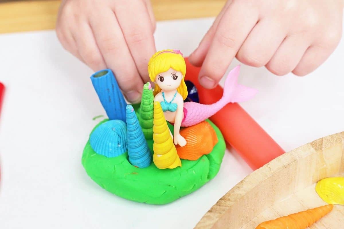 Child uses a rolling pin while creating a mermaid playdough scene with a blonde mermaid figurine, colorful shells, and textured sea themed pieces pressed into green playdough. The hands on activity encourages imaginative play and fine motor skill development through open ended sensory exploration.