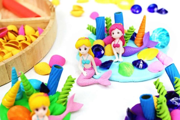 Alt text: Colorful mermaid playdough activity featuring miniature mermaid figurines surrounded by blue playdough, shells, gems, and sea themed loose parts. Children can use the sensory materials to create underwater scenes and imaginative ocean habitats.