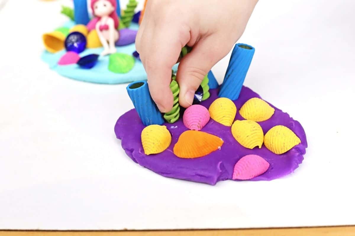Child presses colorful shells, gems, and sea themed pieces into purple playdough during a mermaid playdough activity. A miniature mermaid scene with bright sensory materials encourages creative storytelling and fine motor practice through hands on play.