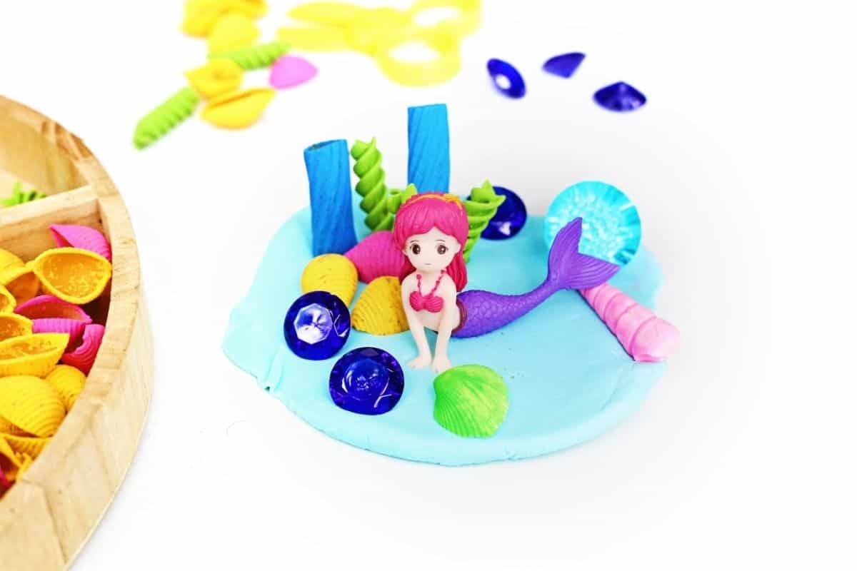 Pink haired mermaid figurine surrounded by colorful shells, blue gems, and sea themed pieces arranged on light blue playdough during a mermaid playdough activity. The underwater inspired sensory play scene encourages imaginative storytelling and creative exploration with ocean themed materials.