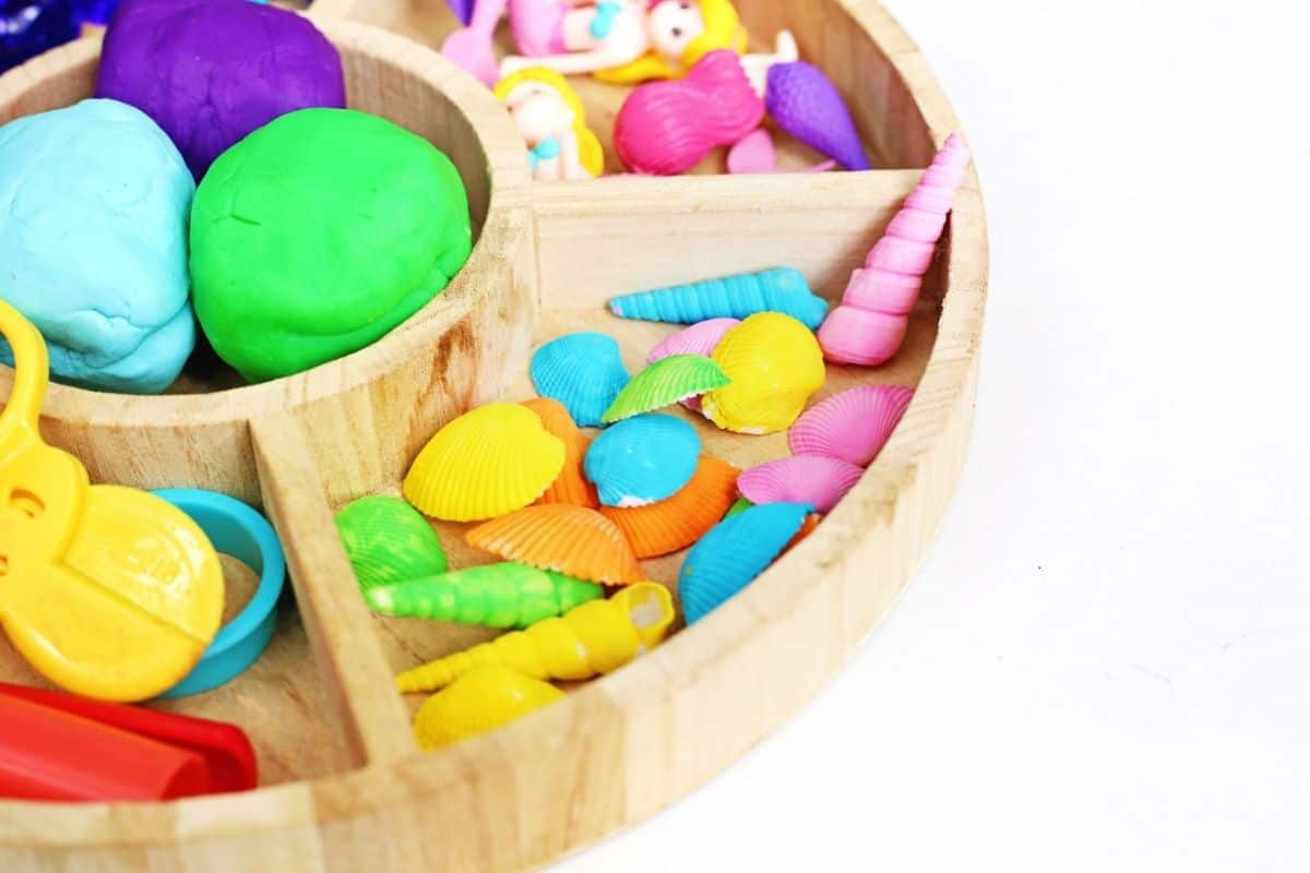 Wooden sorting tray set up for a mermaid playdough activity with colorful playdough, seashells, spiral shells, mermaid figurines, toy scissors, and playdough tools arranged in separate compartments. The organized sensory play materials make it easy for children to create imaginative underwater scenes and explore different textures and colors.