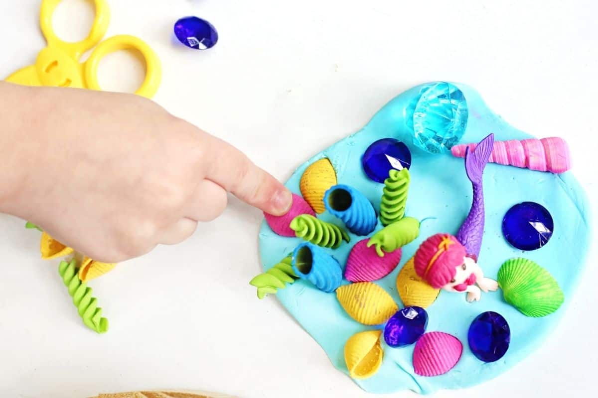 Child points to colorful shells and sea themed pieces arranged around a pink haired mermaid figurine on blue playdough during a mermaid playdough activity. Bright gems, textured materials, and ocean inspired loose parts create an engaging sensory play scene that encourages imaginative storytelling.