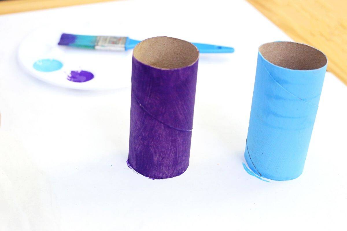 Blue and purple painted toilet paper rolls drying on a white surface before being turned into an octopus toilet paper roll craft. Paint and a brush sit nearby in the background.