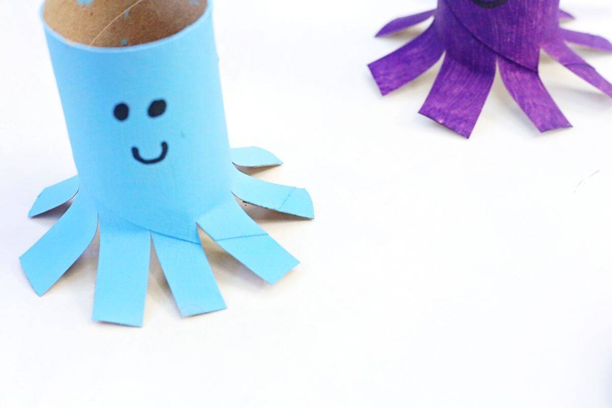 Blue octopus toilet paper roll craft with a smiling face and eight cut tentacles displayed on a white surface. A purple octopus craft appears partially in the background, showing another variation of this easy recycled craft project.