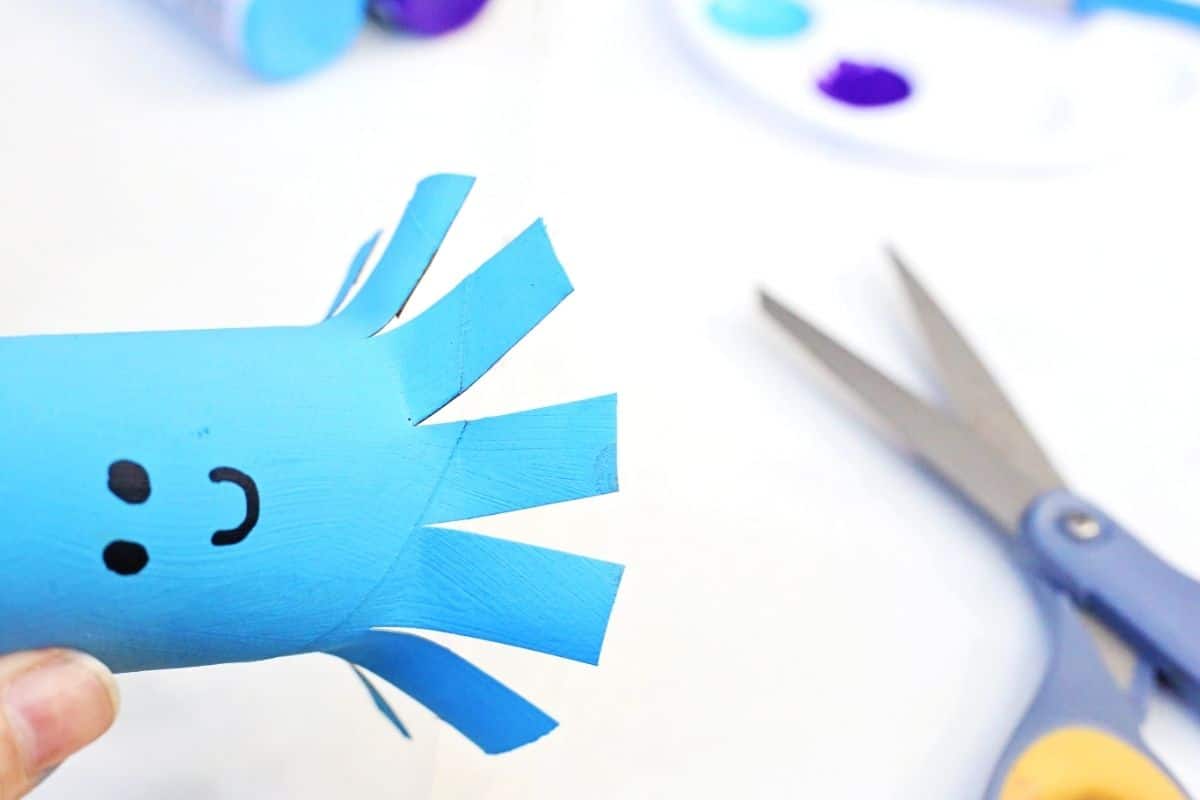Hand holding a blue painted toilet paper roll while scissors are used to cut strips that become tentacles for an octopus toilet paper roll craft. Paint supplies are blurred in the background.