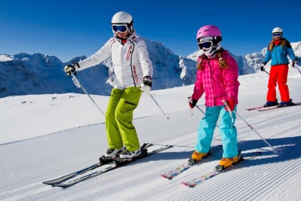 Three skiers wearing colorful winter clothing and helmets ski down a groomed snowy slope under a clear blue sky. Snow covered mountain peaks stretch across the background as the group descends together on a sunny day