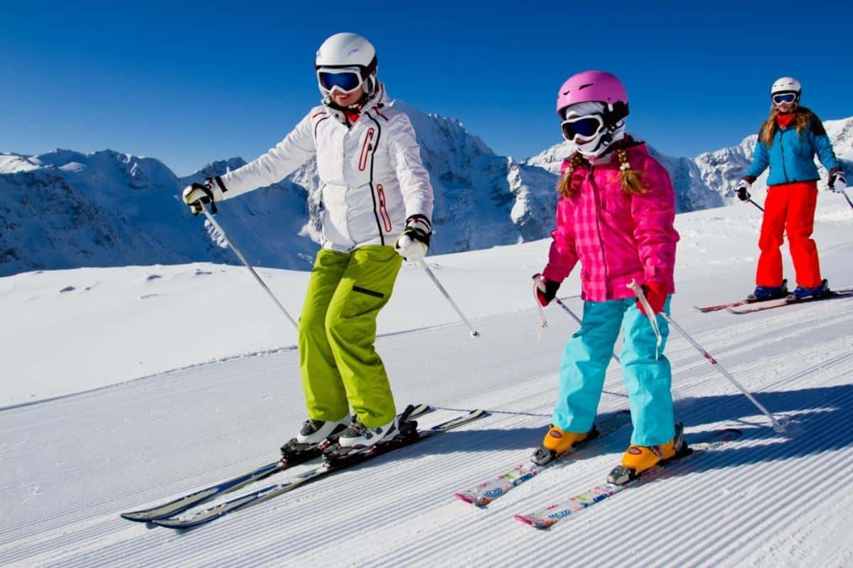 Three skiers wearing colorful winter clothing and helmets ski down a groomed snowy slope under a clear blue sky. Snow covered mountain peaks stretch across the background as the group descends together on a sunny day