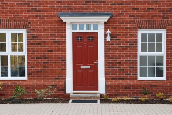 Red composite front door centered on a red brick house exterior with white trim, two small glass panels near the top, and matching white framed windows on either side. The entrance features a small step, exterior wall light, and landscaped border along the front of the property.
