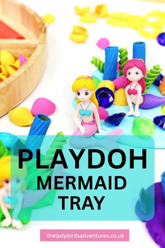 Two small mermaid figures sit in a colorful playdough sensory tray surrounded by bright shells, sea plants, and gem shaped treasures. Text on the image reads "PLAYDOH MERMAID TRAY" and "theladybirdsadventures.co.uk".