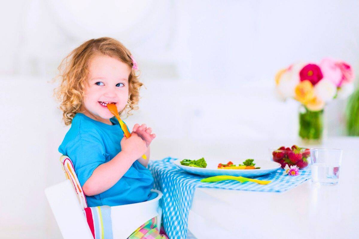 Smiling young child sitting in a high chair at a table with a plate of colorful vegetables, fresh strawberries, and a glass of water while holding a spoon. The child appears happy and engaged at mealtime, illustrating healthy eating for kids in a bright and welcoming home setting.