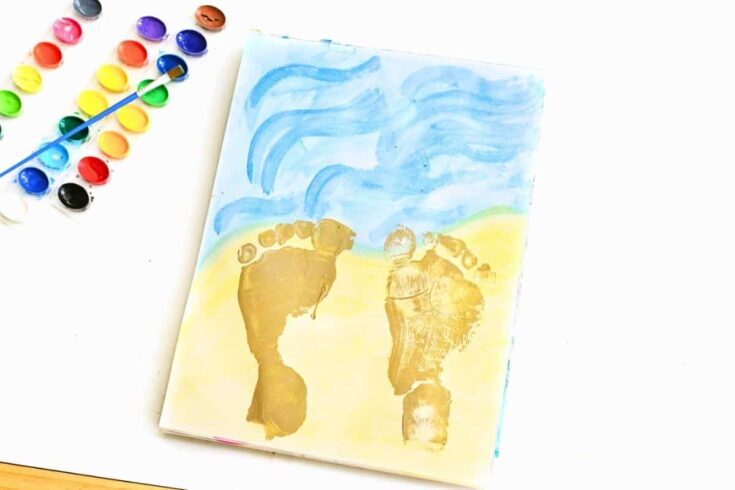 Child footprint prints painted in sandy beige on watercolor paper with soft blue waves and pale yellow sand creating a beach footprint painting. A watercolor paint palette and paintbrush sit beside the finished artwork, showing the materials used to make the beach themed keepsake craft.