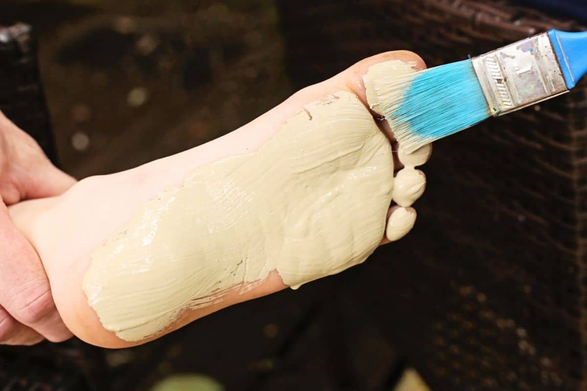 A child’s foot is coated with sandy beige paint using a wide paintbrush as part of a beach footprint painting craft. The paint is brushed evenly across the sole and toes to create a footprint print that resembles footprints left in the sand.