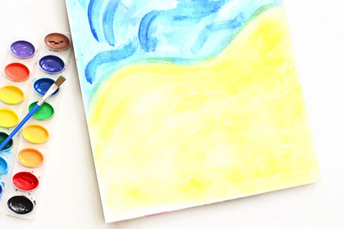 Watercolor beach scene painted with soft blue ocean waves meeting pale yellow sand on a sheet of paper for a beach footprint painting craft. A paintbrush and watercolor palette sit beside the artwork, showing the seaside background before footprints are added.