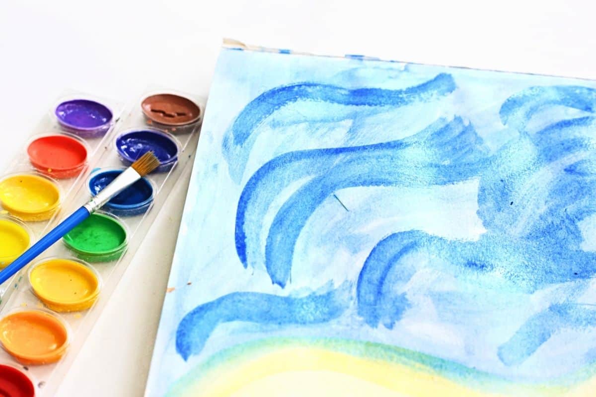 Close up of a beach footprint painting background with layered blue watercolor waves rolling across the top of the page and pale yellow sand below. A watercolor paint palette and paintbrush rest beside the paper, showing the ocean scene being created before footprint prints are added.