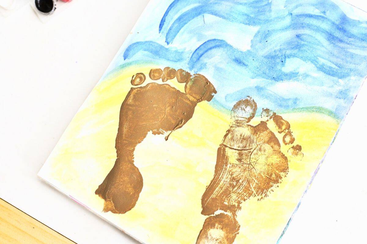 Two sandy beige footprint prints stamped onto a watercolor beach scene with blue ocean waves and pale yellow sand, creating a beach footprint painting keepsake. The footprints appear as if they were left along the shoreline, adding a playful beach themed touch to the handmade artwork.