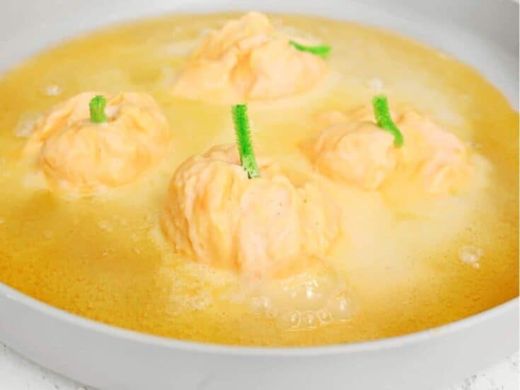 Four small orange pumpkins with green pipe cleaner stems sit in a shallow dish of yellow-orange liquid, surrounded by foam and clusters of bubbles as the fizzy pumpkins experiment reacts.