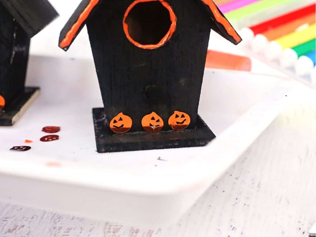 A black wooden birdhouse with bright orange trim around the roof and entrance hole, decorated with three orange jack o lanterns along the base. Colorful craft markers and painting supplies are visible in the background.