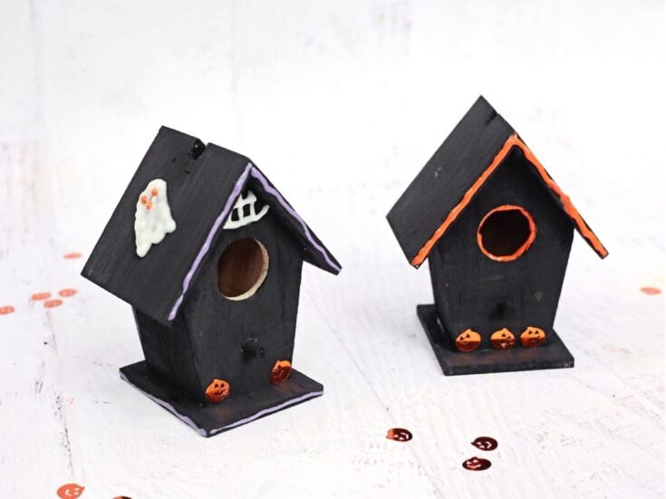 Two finished black wooden birdhouses decorated for Halloween, with purple and orange trim, white ghost details, and small orange jack o lanterns. The spooky designs show the completed haunted birdhouse craft on a white tabletop.