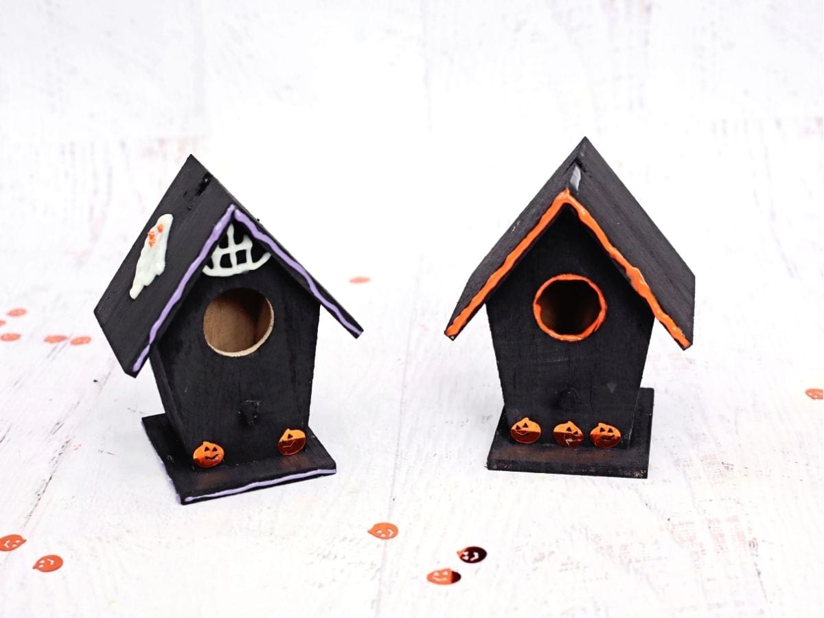 Two finished black wooden birdhouses decorated with purple and orange trim, white ghost details, and small orange jack o lanterns. The spooky birdhouses are displayed side by side as completed Halloween crafts.