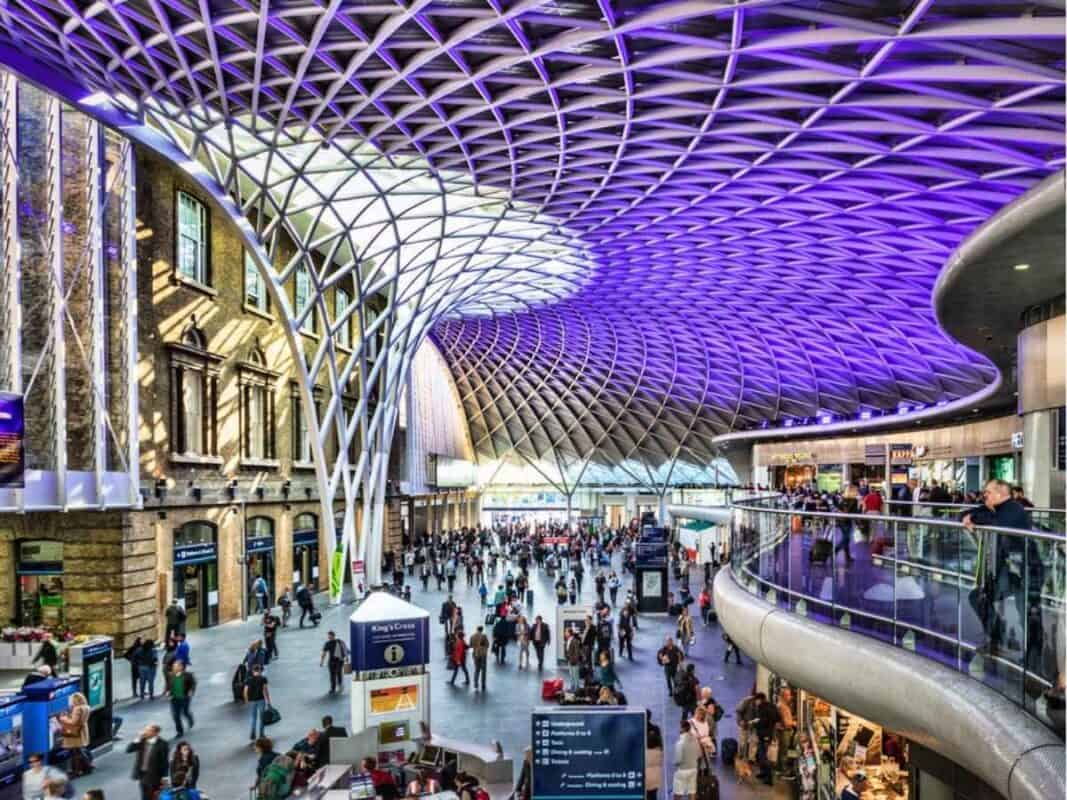 The busy concourse of London Kings Cross station filled with travelers beneath a sweeping geometric steel and glass roof illuminated with purple lighting. The historic brick station facade and clearly marked Underground and Platform signs are visible among the shops and crowds.