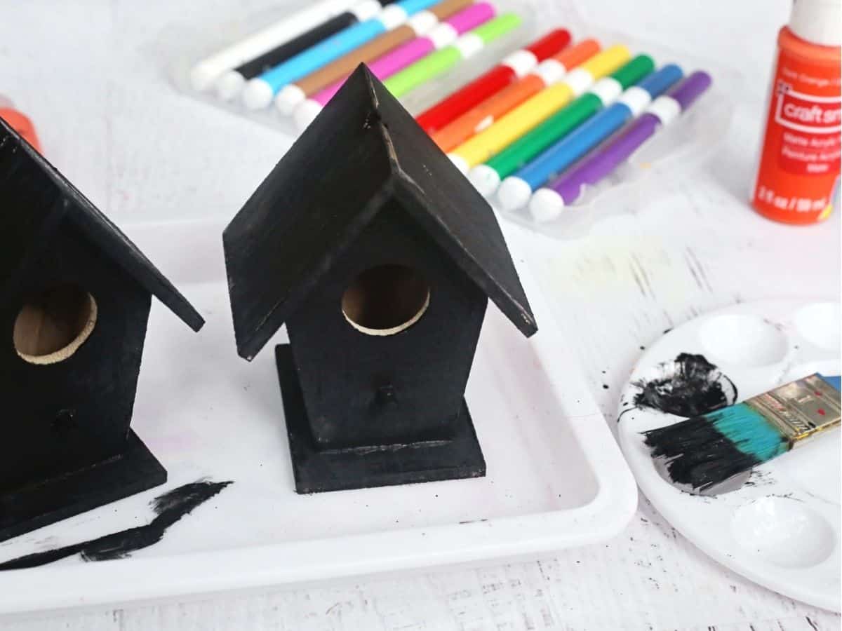 Two wooden birdhouses painted black, with colorful markers, black acrylic paint, brushes, and paint palettes arranged around them. This step shows the birdhouses being painted as part of a haunted birdhouse craft.