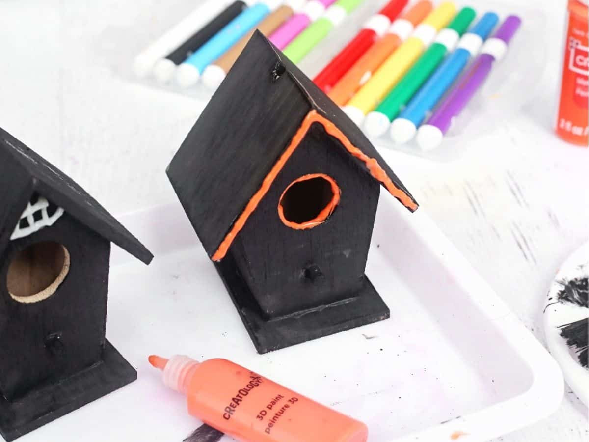 A black wooden birdhouse with bright orange trim around the roof and entrance hole, shown beside another black birdhouse and colorful craft markers. An orange 3D paint bottle is in the foreground as the birdhouses are decorated for a haunted birdhouse craft.