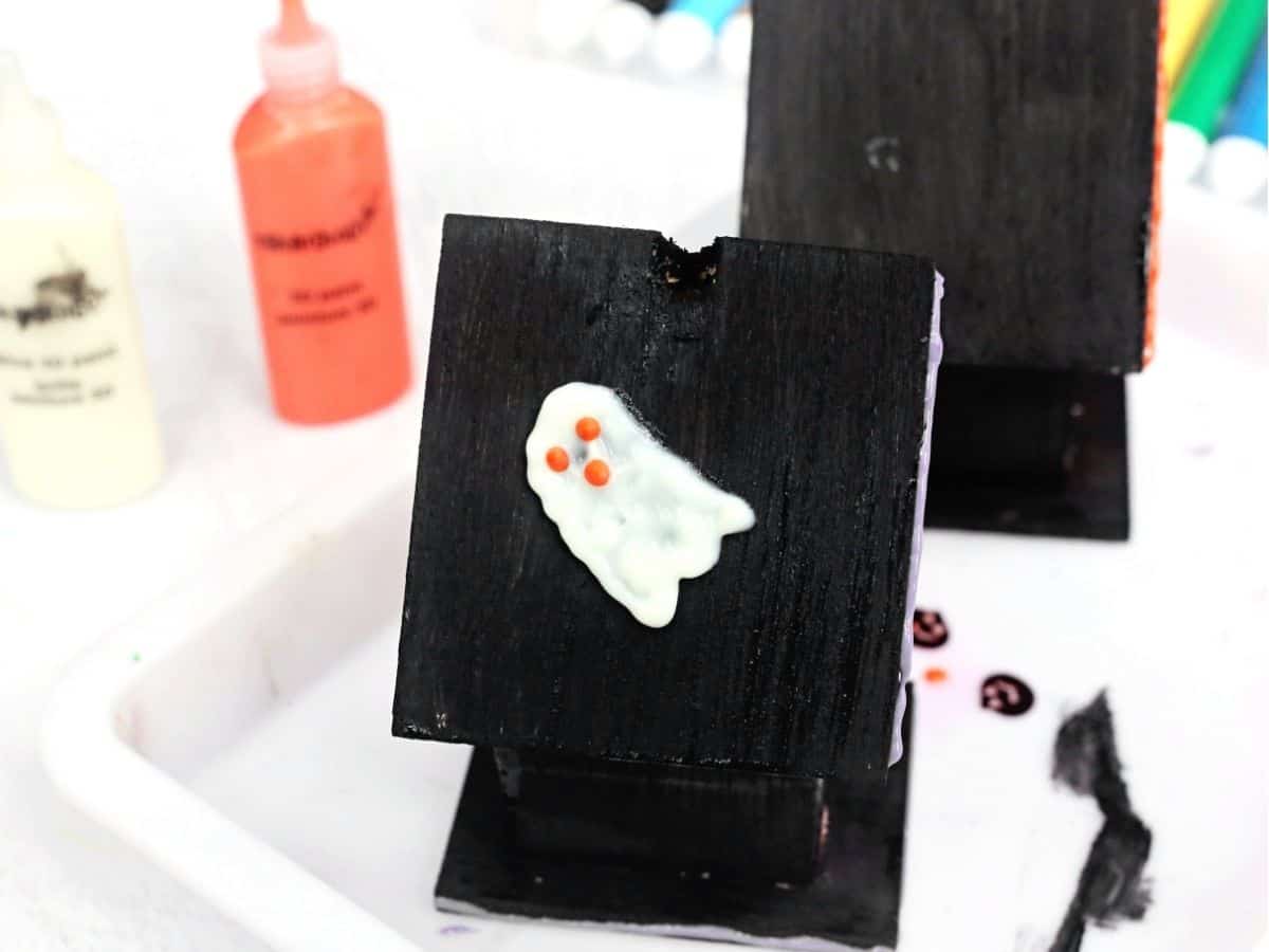 A black wooden birdhouse with a raised white ghost decoration featuring small orange details on its side. Paint bottles, a palette, and brushes surround the birdhouse as it is decorated for a haunted birdhouse craft.