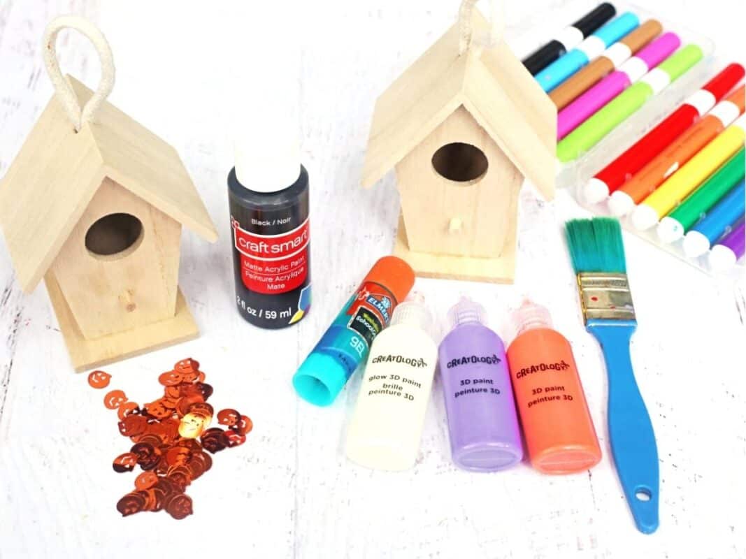 Two unfinished wooden birdhouses surrounded by black acrylic paint, colorful markers, 3D paint bottles, orange glue, a paintbrush, and orange jack o lantern embellishments. These supplies are used to make a haunted birdhouse craft for Halloween.